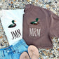 A styled flat-lay of two graphic t-shirts with duck illustrations and pocket monograms, paired with light blue denim shorts and beige flip-flops on a bed of small gray pebbles. One white t-shirt has a mallard graphic above the text 'JMN', and one brown t-shirt has a mallard graphic above the text 'MRM'.
