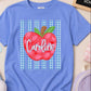 A personalized blue short-sleeve t-shirt with a red apple graphic and the name 'Caroline' printed on the front, styled in a flat lay with school accessories.