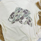 Ivory tee with a camouflage fish design on a rug surface.