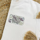 A white t-shirt with camouflage graphics and the text 'DAD' on the front pocket, displayed on a woven surface with beaded decorations in the background.