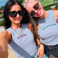 Two women wearing gray tank tops with 'TIDE' and 'Hornets' text, standing outdoors near a sports field.
