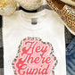 White t-shirt with 'Hey There Cupid' design, leopard print shoes, and candle on a checkered fabric background