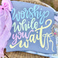 Decorative sign with 'Worship while you wait' text on a blue background, surrounded by decorative elements.
