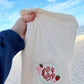 Beige fabric with a monogram and strawberry design held by a hand against a beach background