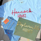 Light blue t-shirt with 'Hancock Hawks' text, Levi's jeans, and green tote bag with 'Poplarville Hornets' text on a wooden surface.