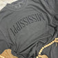 Gray t-shirt with 'MISSISSIPPI' printed on it, draped over a textured surface.