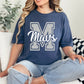 Woman wearing a navy blue t-shirt with 'Mavs' printed on it, sitting on a couch.