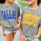 Two women walking outdoors wearing matching Mossy Oak Bottomland graphic t-shirts. The woman on the left has a blue 'DALLAS' print and the woman on the right has a yellow 'TENNESSEE' print, both paired with denim shorts and sunglasses.