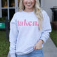 A woman standing outside wearing a gray sweatshirt with the text 'taken' printed in pink, paired with blue denim shorts with a frayed hem.